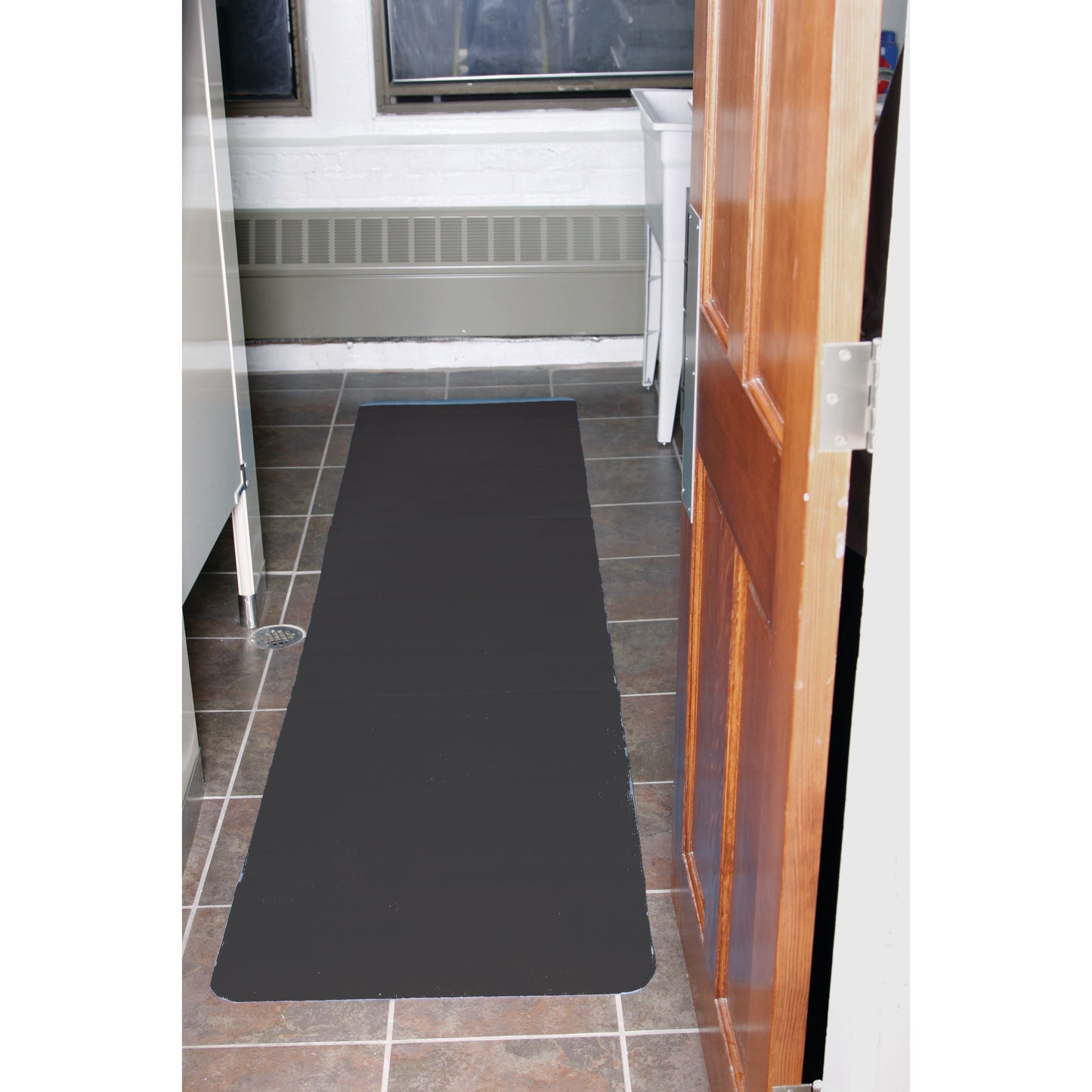 A long, black AliMed Safety NoSlip Mat is placed on a tiled floor in a room with a wooden door and a white sink.