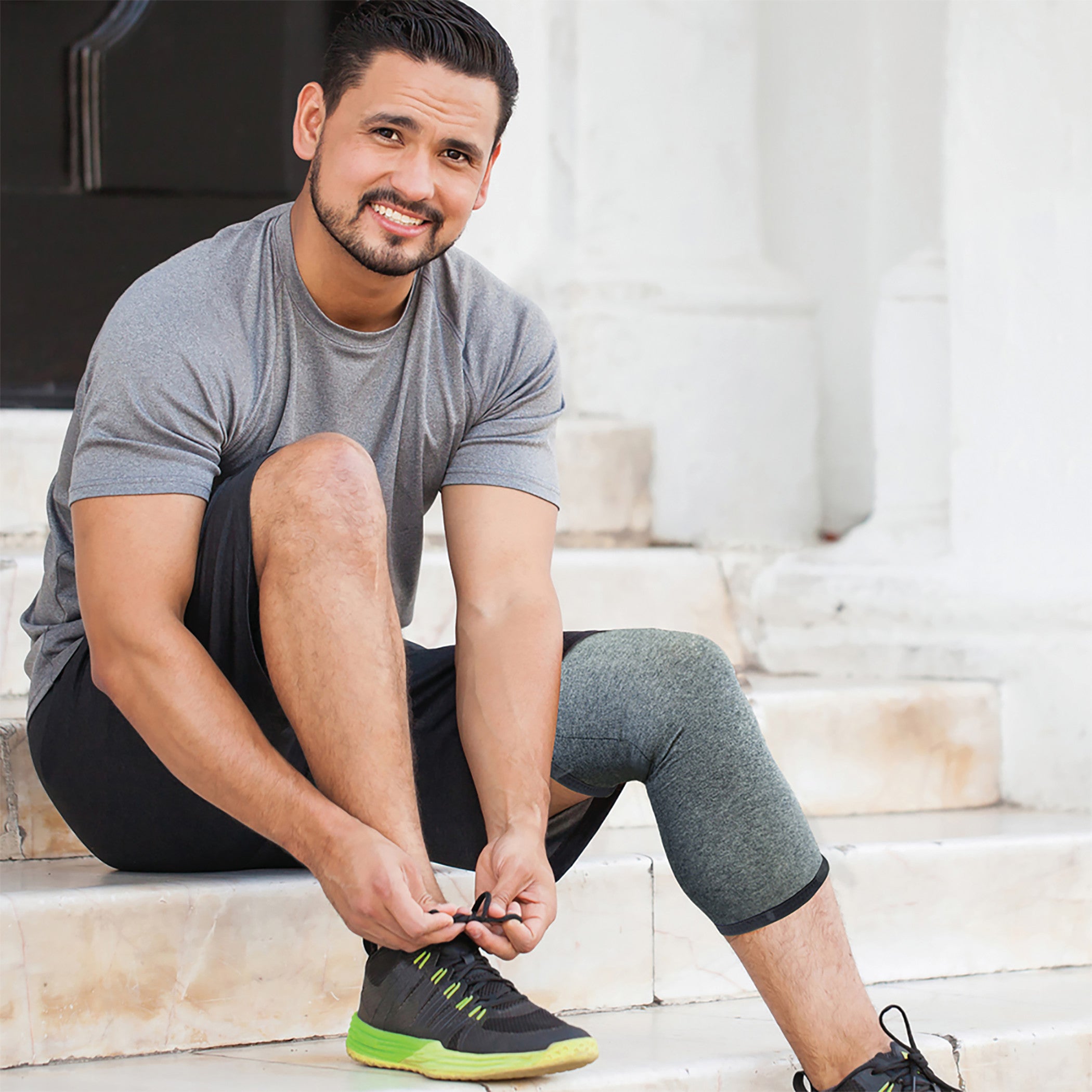 A man ties his shoe while wearing the Brownmed IMAK Compression Arthritis Knee Sleeve on his right knee, seated on steps.