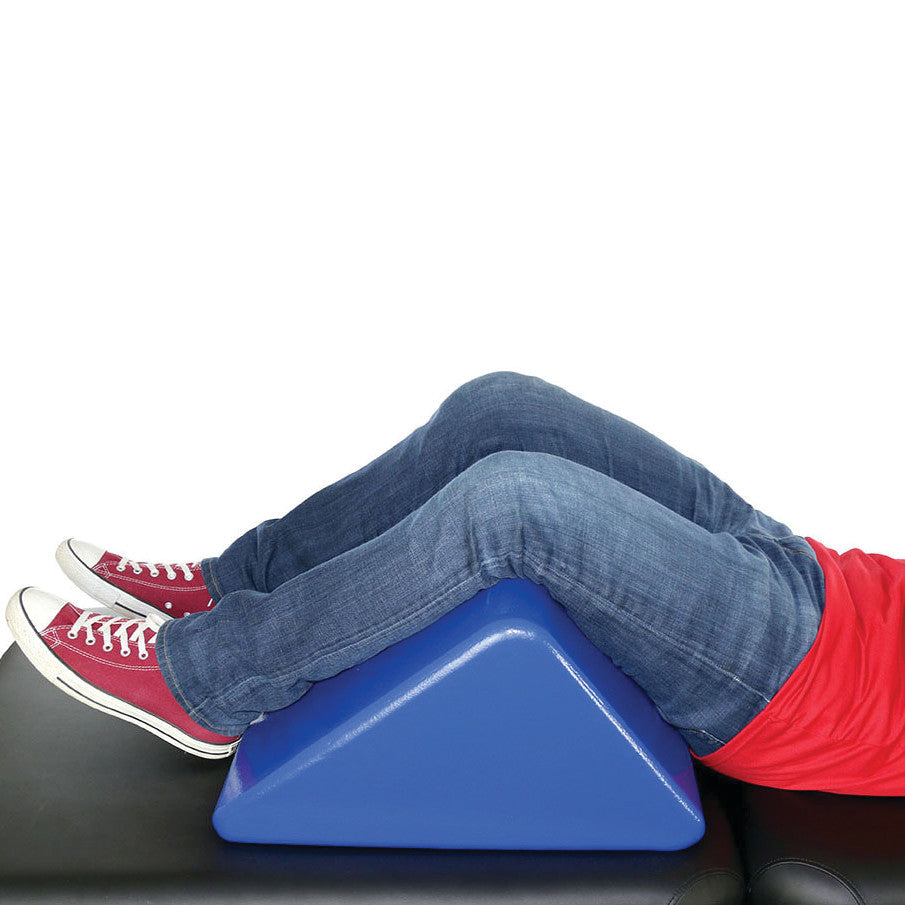 Person lying back with legs bent over a blue Skillbuilders Wedge Knee Bolster on a black surface, wearing jeans and red sneakers.