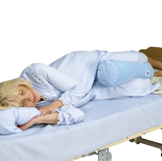 A person sleeps on their side with a SkiL-Care Ultra-Soft Bed Bolster placed between their knees on a blue-sheeted bed.