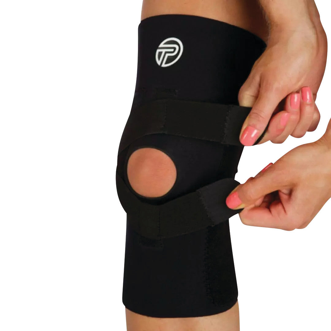 A person adjusts the J-Lat Knee Support, a black brace with an open patella design and adjustable straps, worn on the knee.