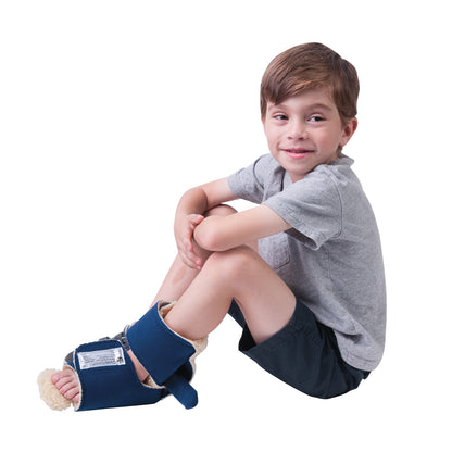 A child wearing a Comfy Splints Pediatric Ambulating Contracture Boot on the foot, sitting cross-legged, smiling, in a grey shirt.