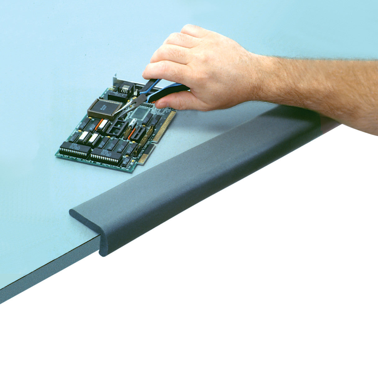 A hand works on a circuit board on a table, protected by an AliMed SoftEdge Edge Protector along the table edge.
