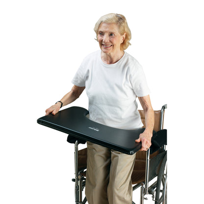 An elderly woman stands holding a SkiL-Care Lift-Away Lap Tray, attached to a black wheelchair, smiling and wearing a white shirt.