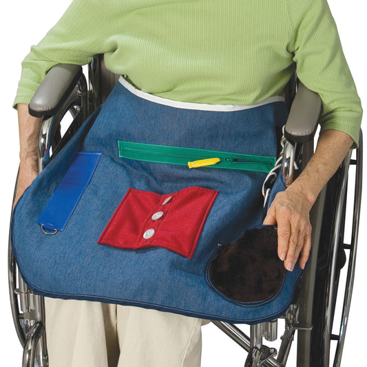A person in a wheelchair wears a Posey Activity Apron. The apron has button, zipper, and pocket activities on a denim fabric.