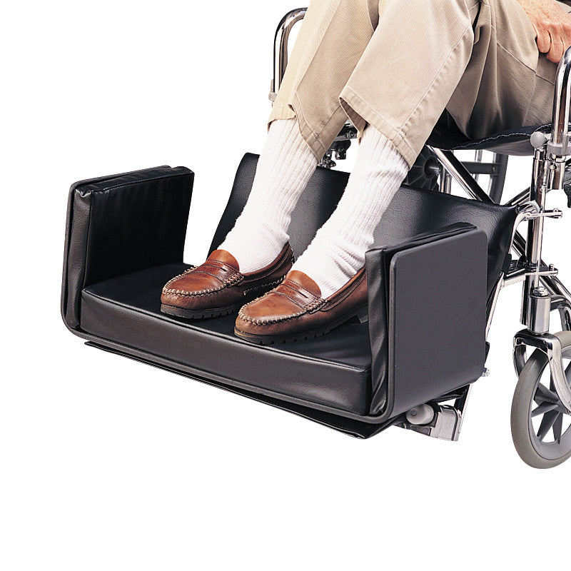Man's legs are resting on a SkiL-Care Side-Kick Pad attached to a wheelchair, with brown shoes and white socks visible.