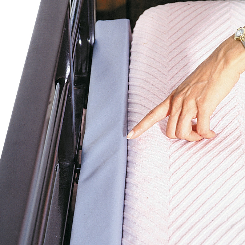 A hand gestures towards the AliMed Bed Stuffer Safety Bolster, placed beside a bed with a pink textured blanket.