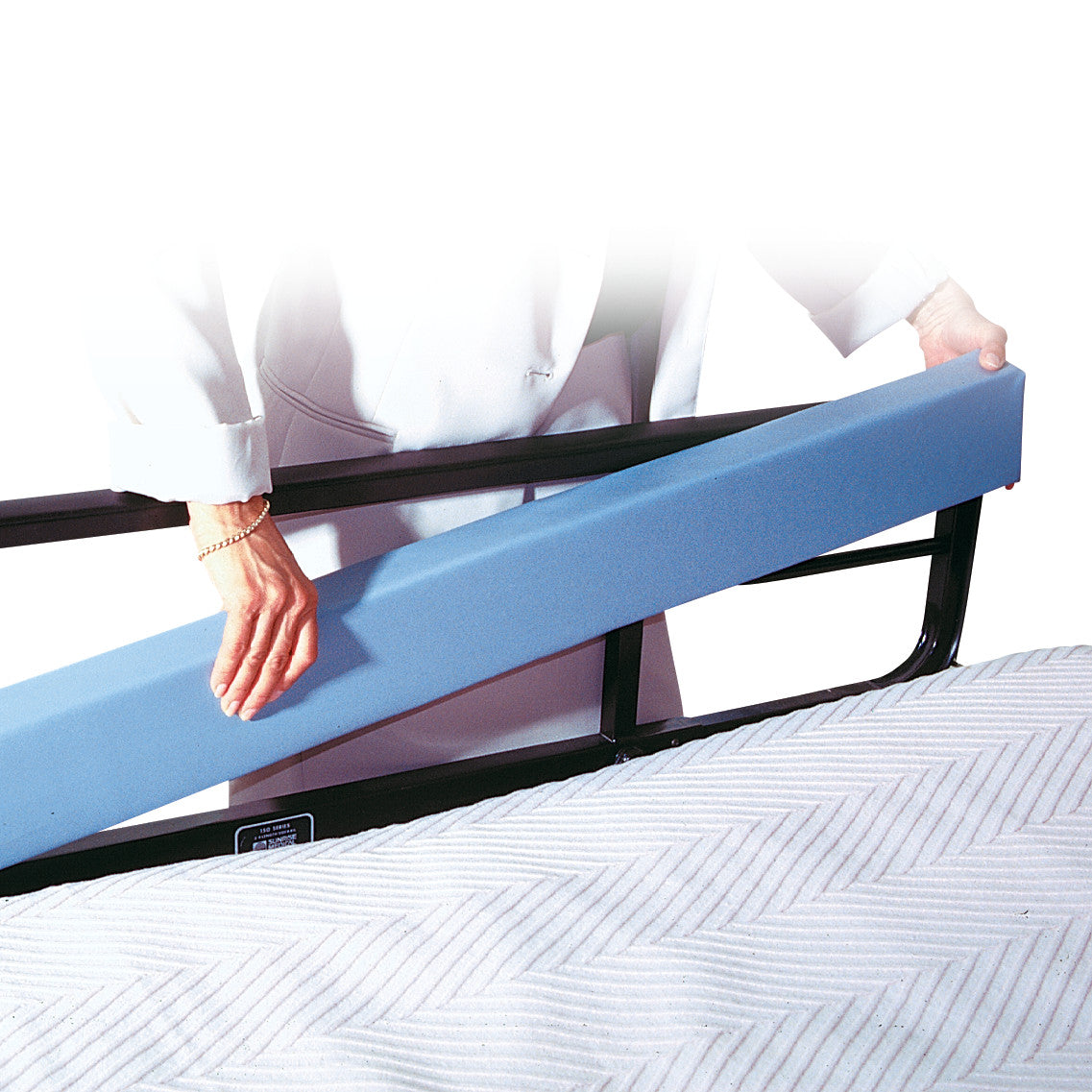 A person places an AliMed Bed Stuffer Safety Bolster, a blue foam cushion, between a bed's rail and mattress for safety.