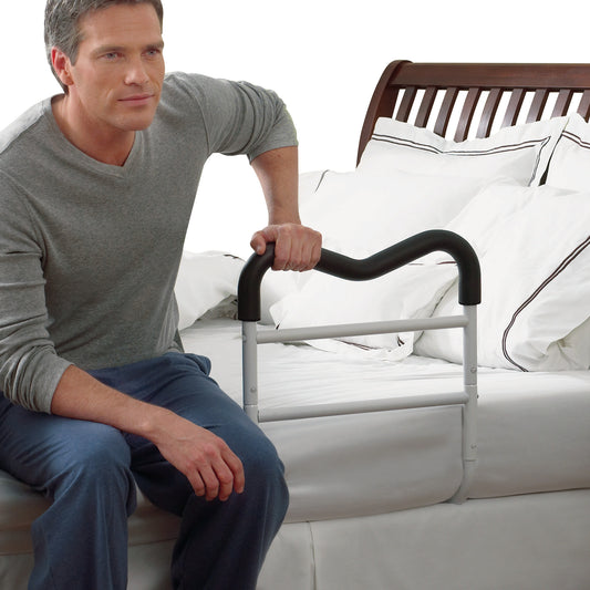 A person uses an M-Rail Assistive Bed Rail, attached to a neatly made bed, for support. The rail is white with a black top handle.