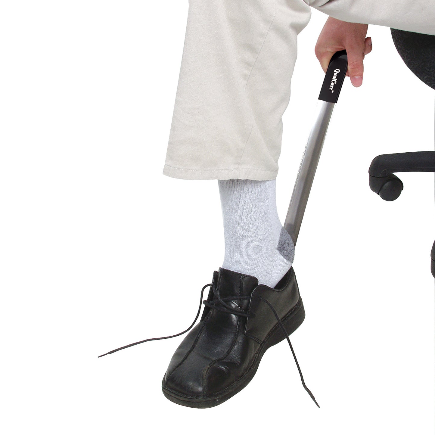 A person uses a Therafin Stainless Steel Shoehorn to put on a black shoe, while seated, wearing light socks and beige pants.