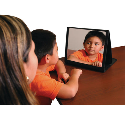A child looks in a tabletop mirror, part of a Therapy Kit, while seated at a wooden table with a woman observing.