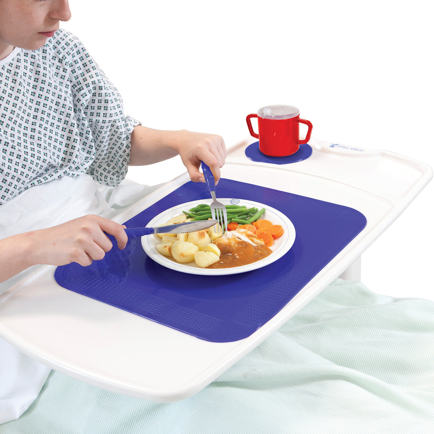 A person in bed uses a Dycem Nonslip Rectangular Matting on a tray. The mat holds a plate of food and a red handled cup.