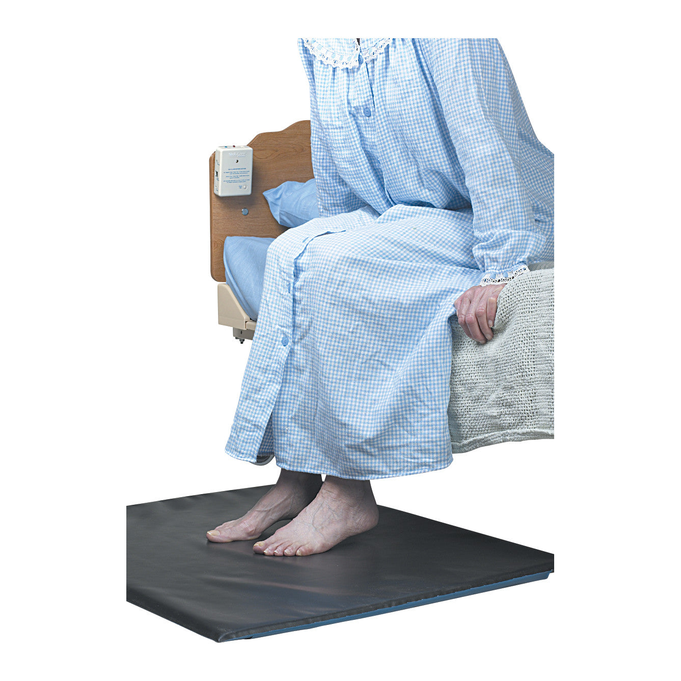 Person sitting on a bed with feet on the SkiL-Care FloorPro Floor Mat Alarm System, wearing a blue checkered nightgown.