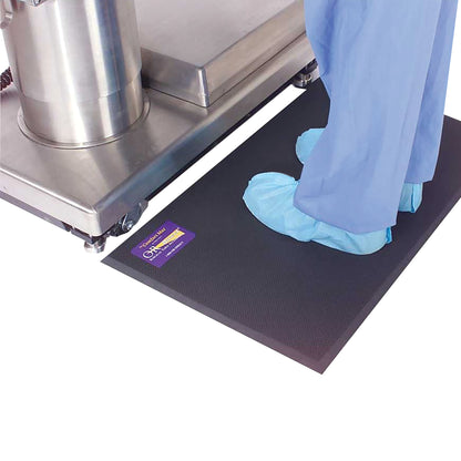 A person wearing blue shoe covers stands on an Allen Comfort Mat beside stainless steel equipment on wheels.