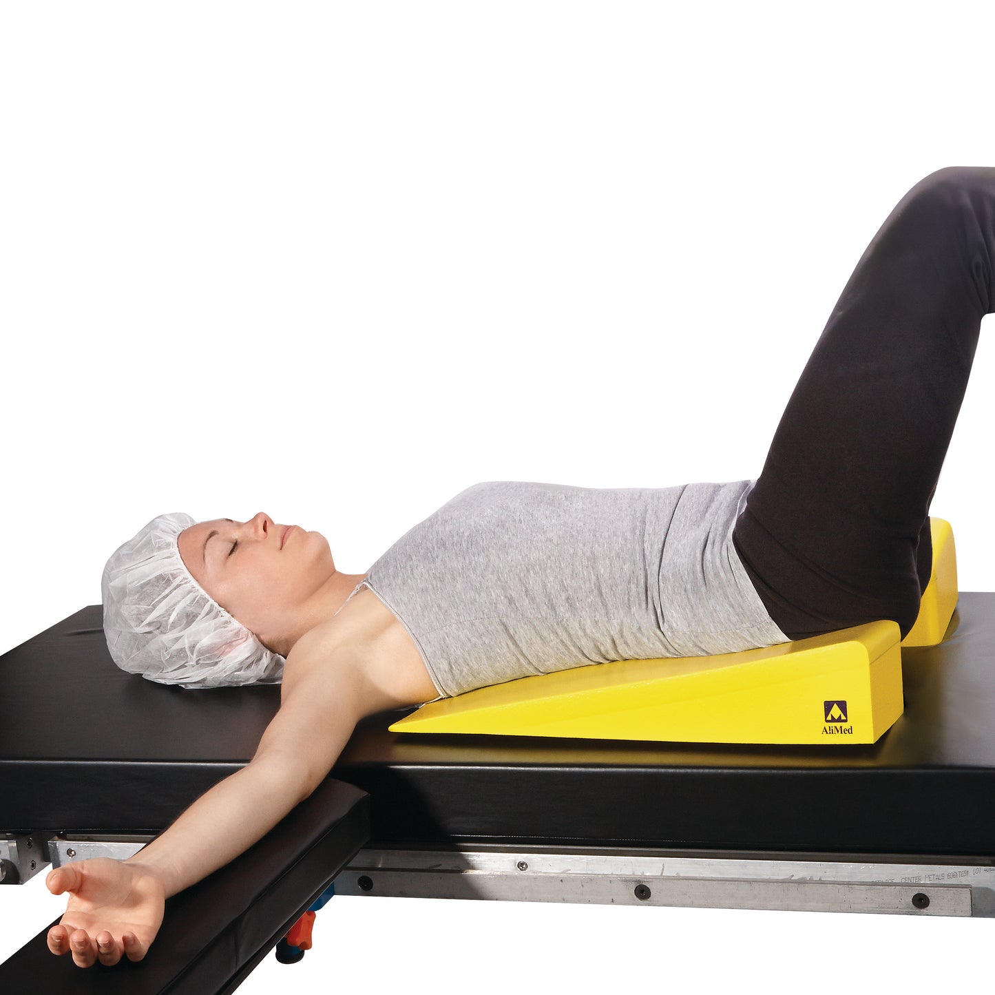Person lying on a padded table using the AliMed Protecta-Coat Perineal Wedge. The wedge is yellow and supports the lower legs.