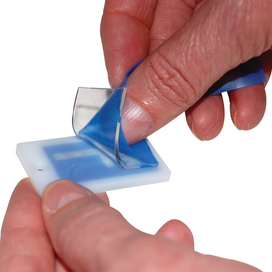 A person peels blue film off a white plastic tag labeled "Marker Mate." The tag has a hole for hanging or attaching.