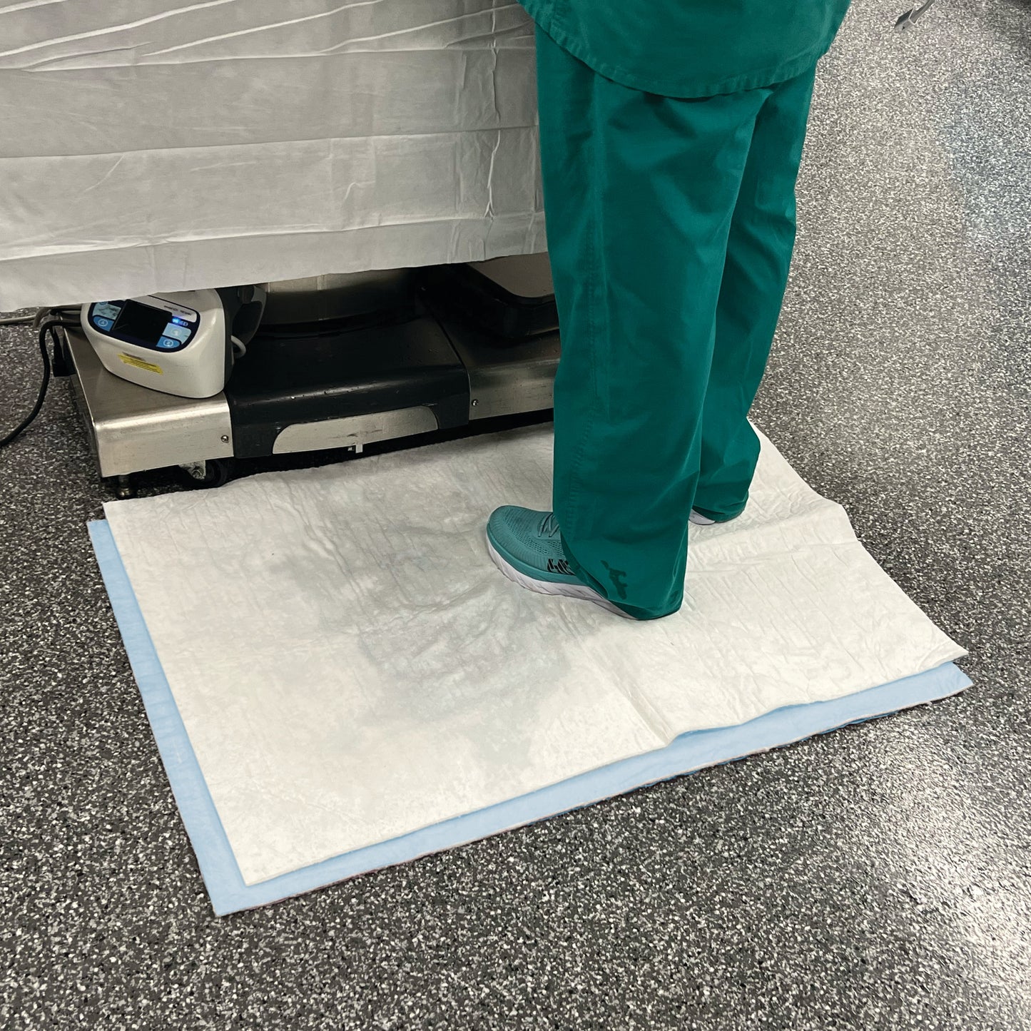 Person standing on a SpilfyterMED White Absorbent Mat for Emergent Use, placed on a speckled floor, beside medical equipment.