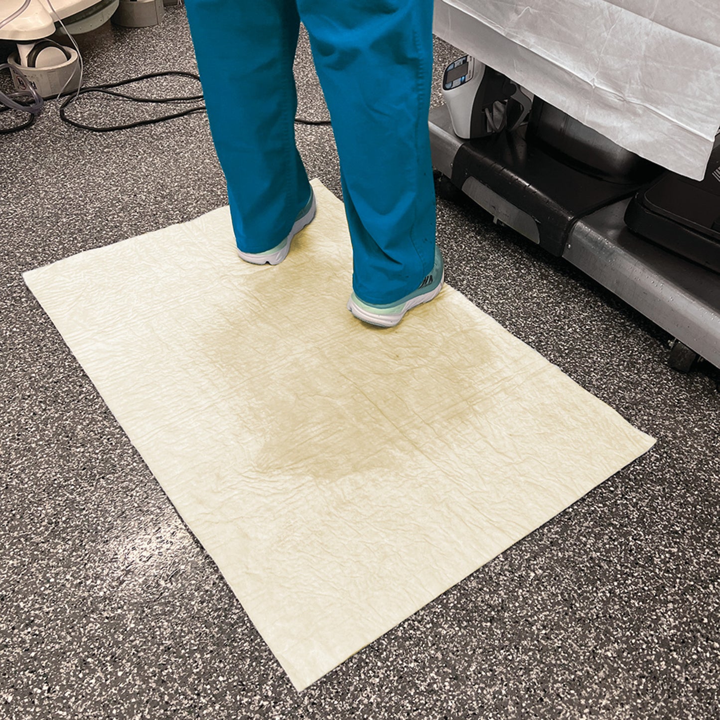 Person standing on a SpilfyterMED Yellow Absorbent Mat for Moderate Spillage, placed on a speckled floor, near medical equipment.