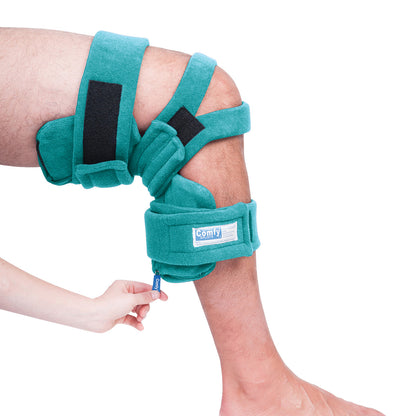 A leg is fitted with a Comfy Splints Pediatric Locking Knee Orthosis, teal in color, secured with straps. A hand adjusts it.