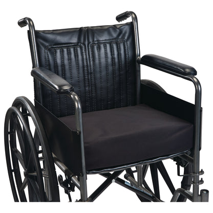 A black SkiL-Care Ultra Gel-Foam Vinyl Wedge Wheelchair Cushion is placed on a wheelchair seat with armrests and large wheels.