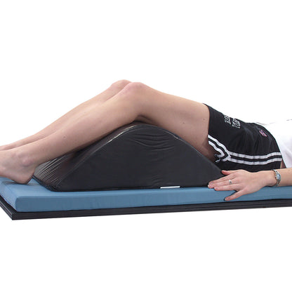 A person uses the AliMed Vinyl-Covered Deluxe Knee Bolster/Lumbar Positioner. Legs rest on the wedge-shaped cushion on a mat.
