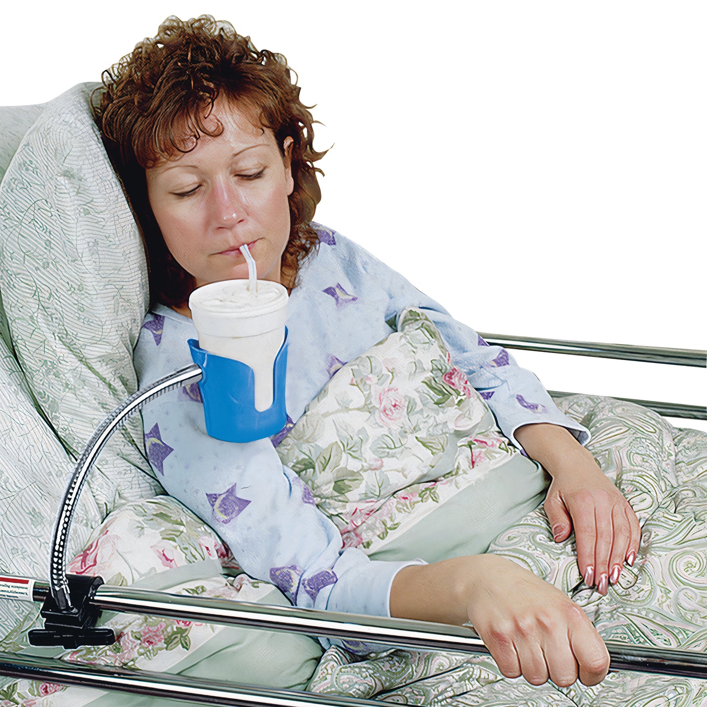 A woman in bed uses a Bedside Beverage Holder to drink from a cup with a straw. The holder is clamped to the bed rail.