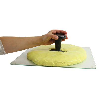 A hand presses a Puttycise Tool Set handle into yellow putty on a flat surface, demonstrating its use for hand therapy exercises.