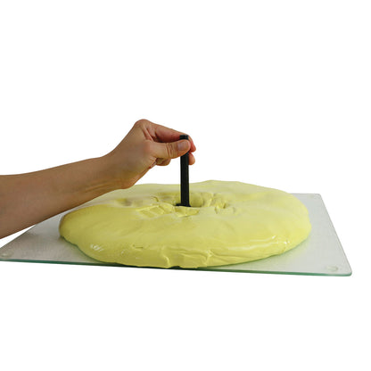 A hand uses a black tool from a Puttycise Tool Set, pressing it into yellow putty on a glass surface.