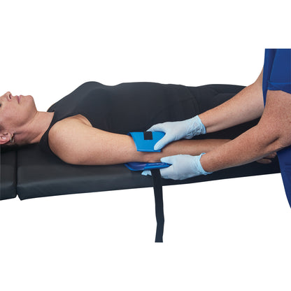 A person lies on a table while another person places the AliMed AliBlue Gel Ulnar Nerve Protector under their arm, securing it with a strap.
