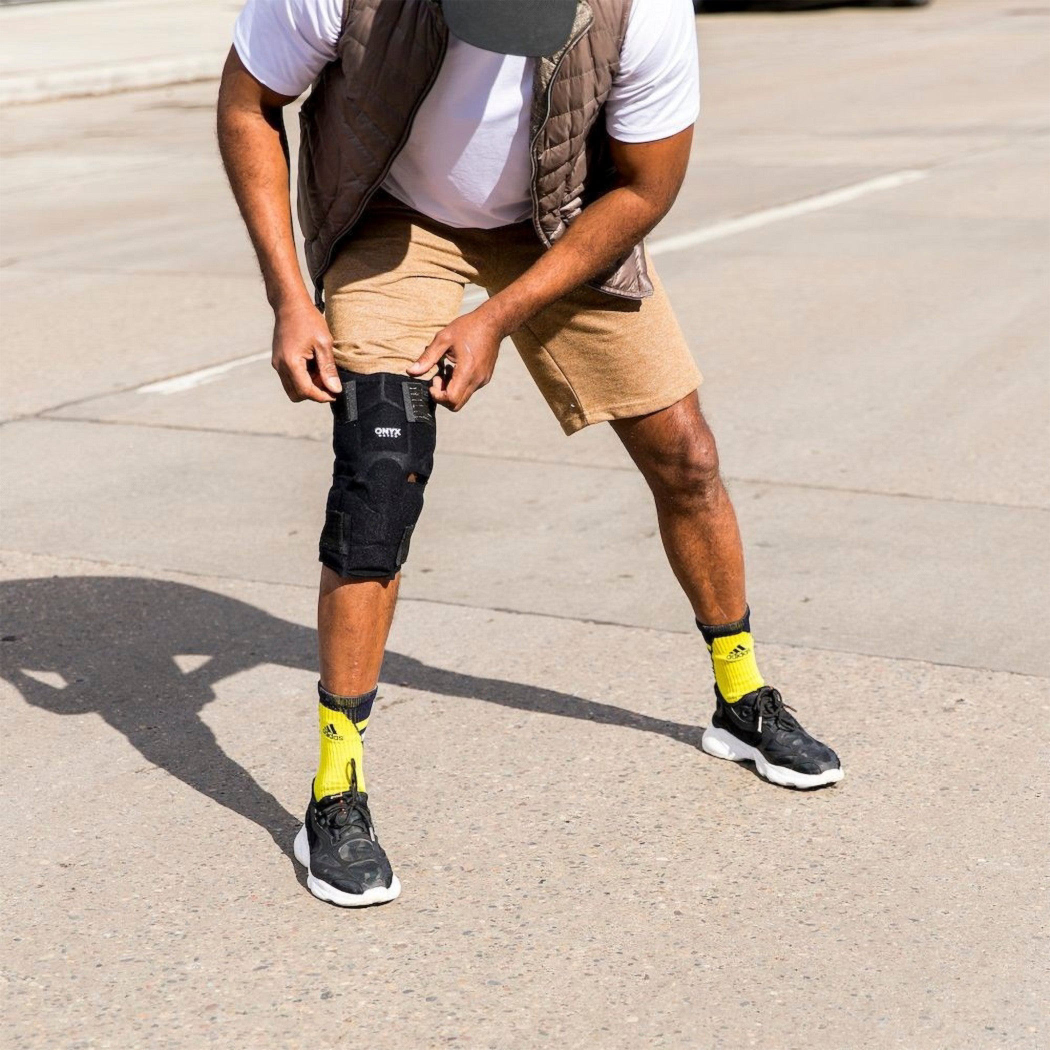 A person adjusts a Xena Therapies Onyx Cool Knee brace outdoors, wearing shorts, a white shirt, and sneakers with bright socks.