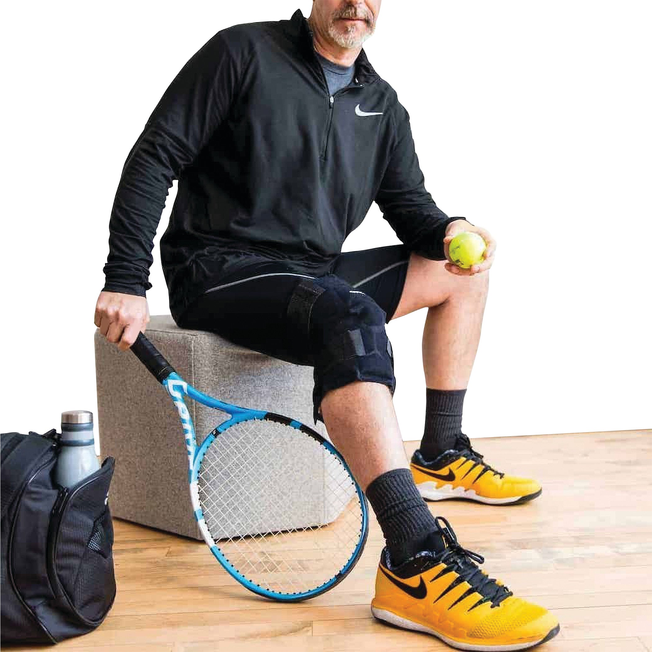 A person is sitting, holding a tennis ball and racquet, wearing the Xena Therapies Onyx Cool Knee brace on their right leg.