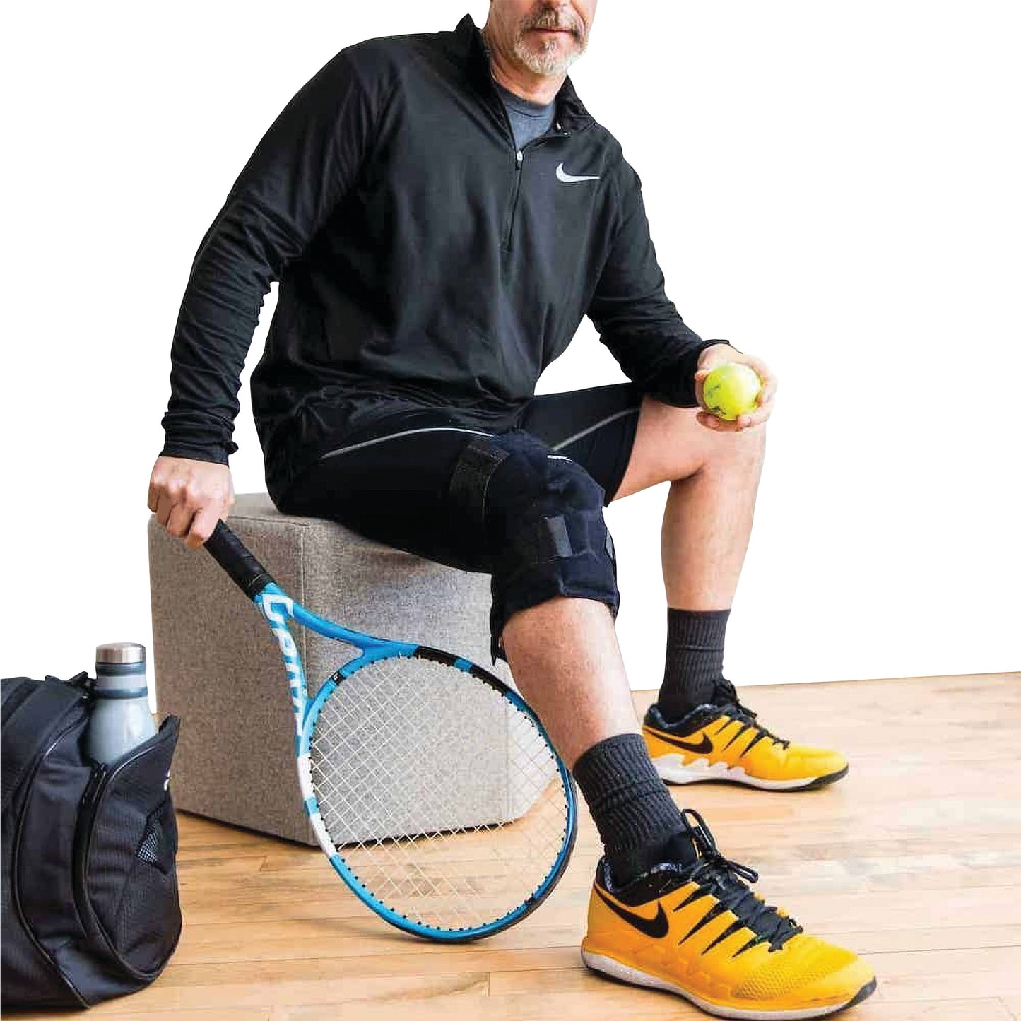 A person is sitting, holding a tennis ball and racquet, wearing the Xena Therapies Onyx Cool Knee brace on their right leg.