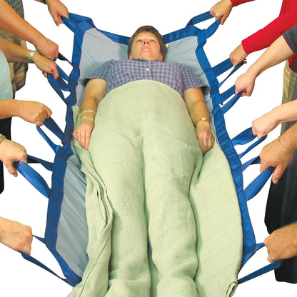 A woman lies on the SkiL-Care Bariatric Turner, held by multiple people using blue handles, with a light green blanket over her.