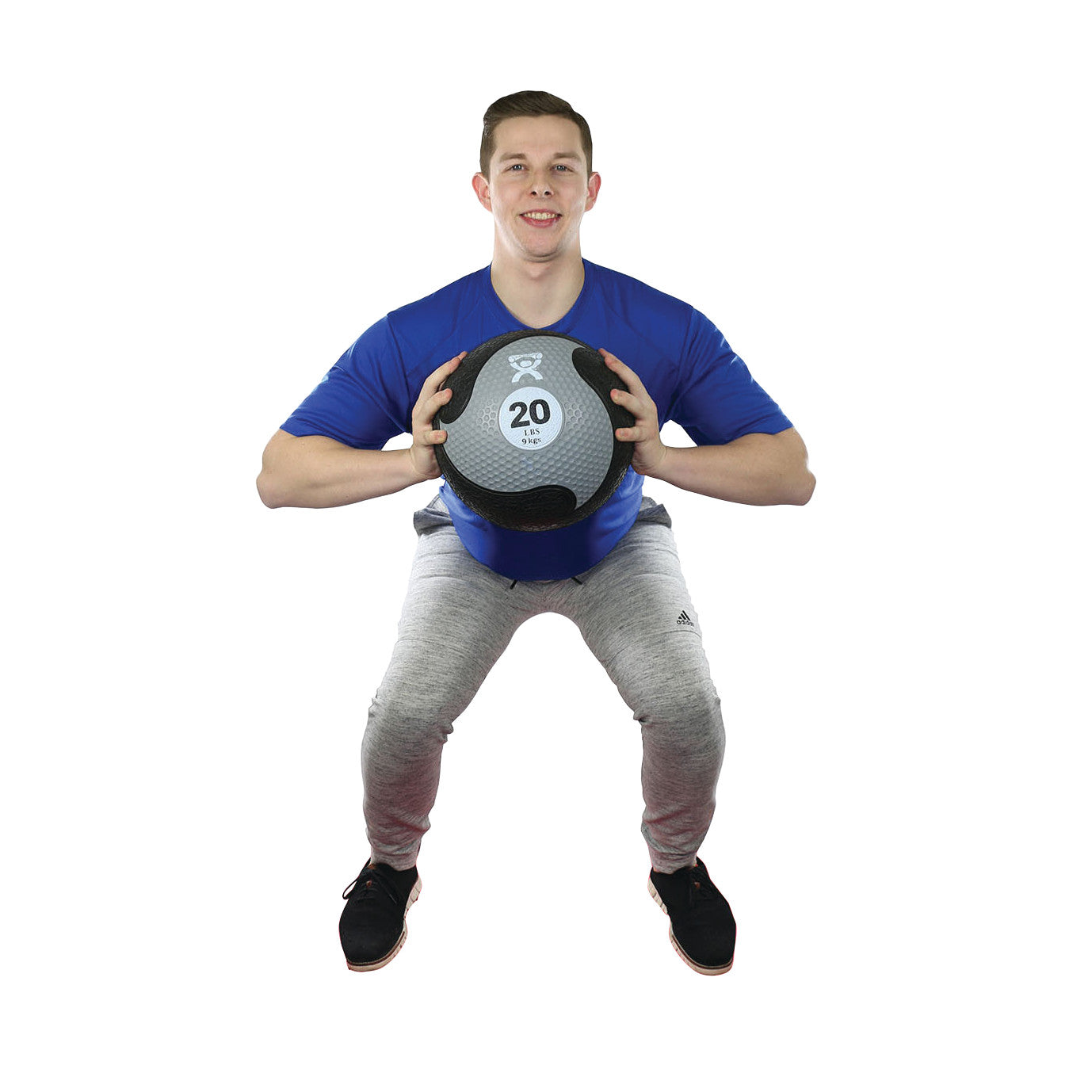 A person squats, holding a CanDo Rubber Medicine Ball weighing 20 lbs, wearing a blue shirt and gray pants.
