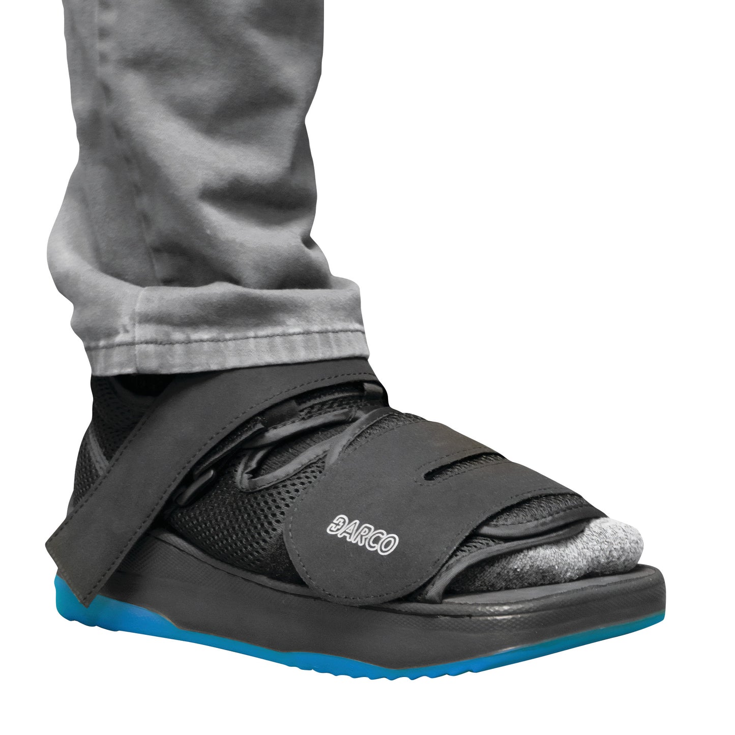 A person wearing the DARCO MedSurg DUO post-op shoe with black mesh, Velcro straps, and a bright blue sole, over light gray pants.