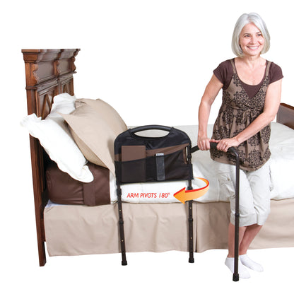 A woman uses a Stander Mobility Bed Rail, which pivots 180�, for support beside a bed. The rail has a storage pouch attached.