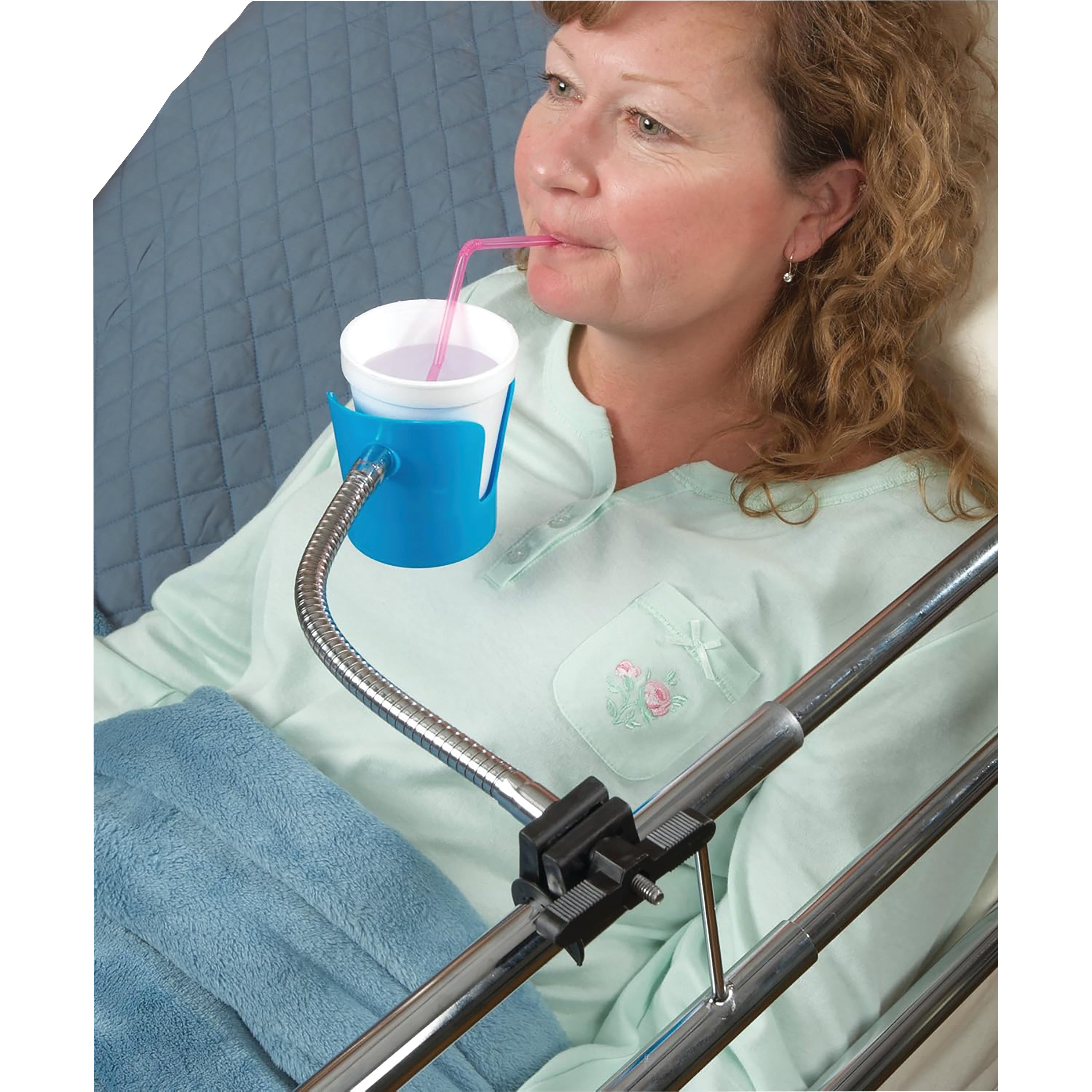A woman is lying in bed using the Bedside Beverage Holder, which clamps to the bed rail and supports a drink with a straw.