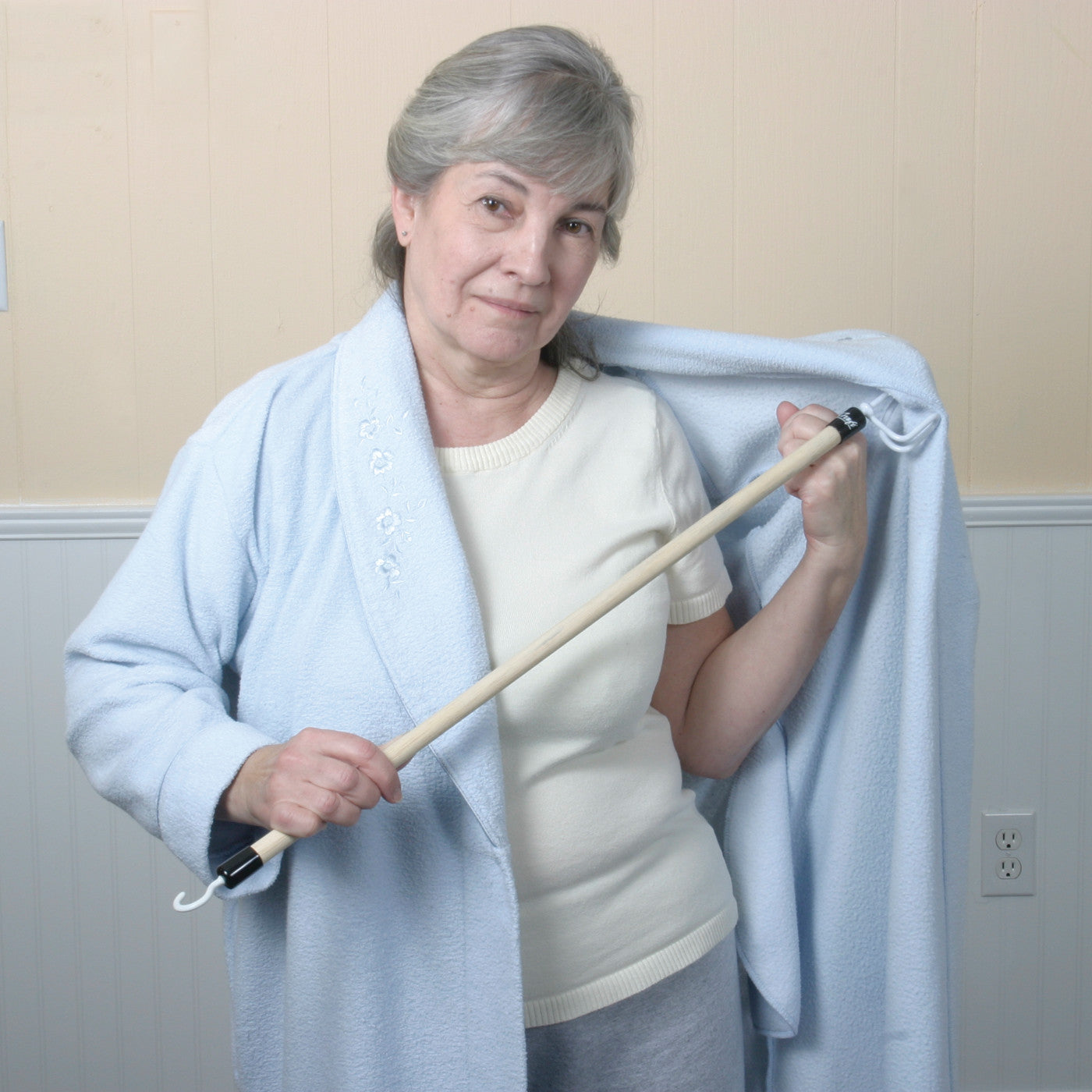 An older woman using the Big Hook Dressing Stick to put on a light blue robe over a white shirt, standing indoors.