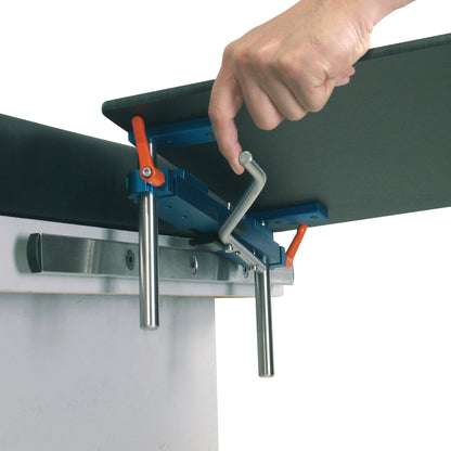 A hand adjusts the AliMed Quick-Snap Table Clamp, made of blue and metal components, securing it to a table edge.