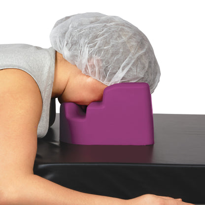 A person wearing a hairnet rests their face on a purple Light Cloud Prone Headrest, which is on a black surface.