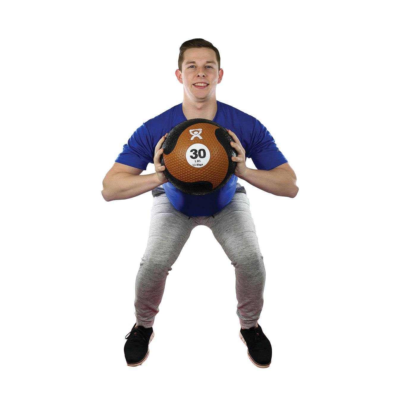 A person squats while holding a 30-pound CanDo Rubber Medicine Ball, wearing a blue shirt and gray pants against a white background.