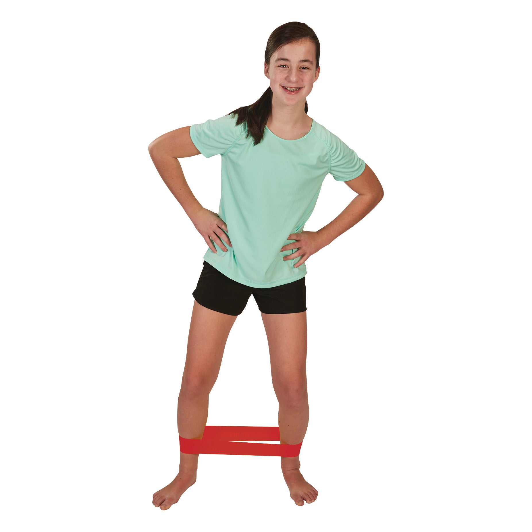 A child stands smiling with a red CanDo Band Exercise Loop around her legs, wearing a light green shirt and black shorts.