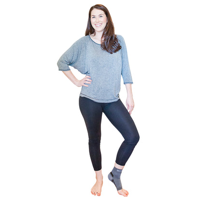 A woman wears the Nice Stretch Total Solution PF Kit on one foot, standing in a relaxed pose with leggings and a long-sleeve top.
