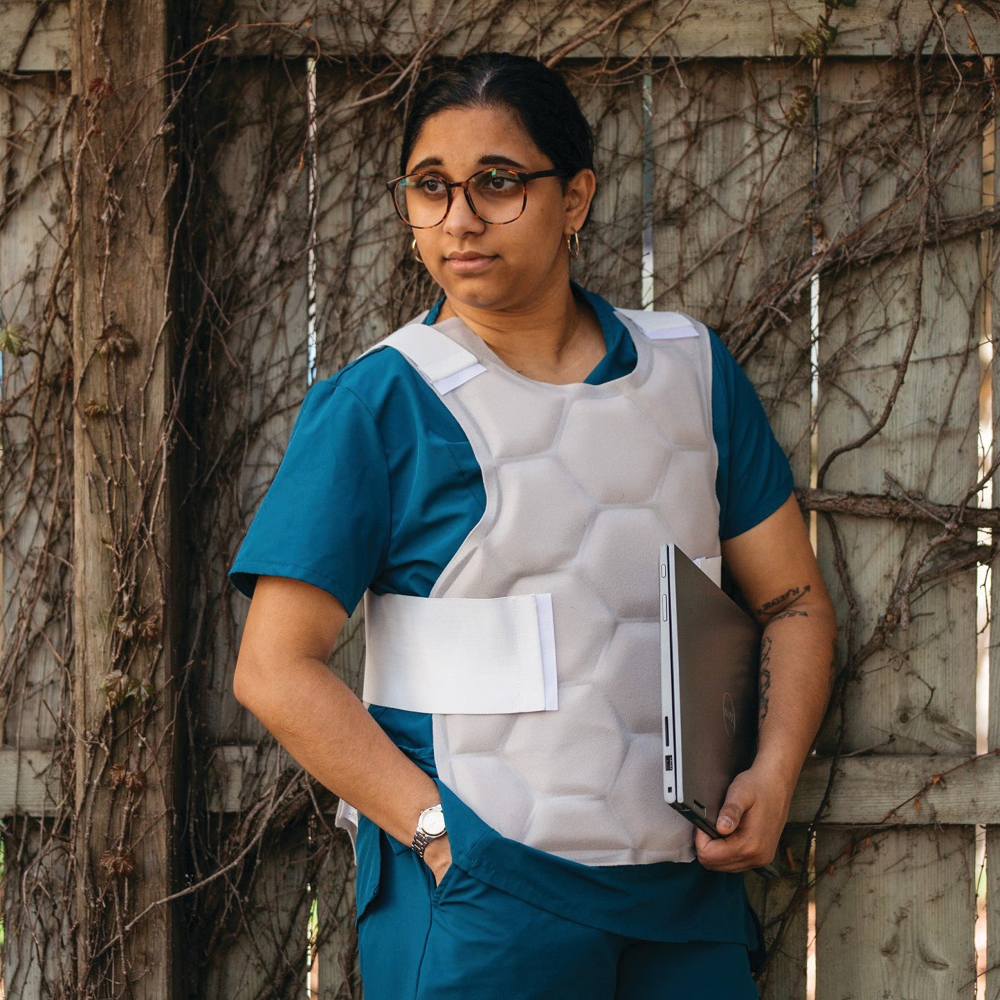 A person wears the Xena Therapies Onyx Cool Safety Pro Vest, holding a laptop, standing against a vine-covered wooden fence.