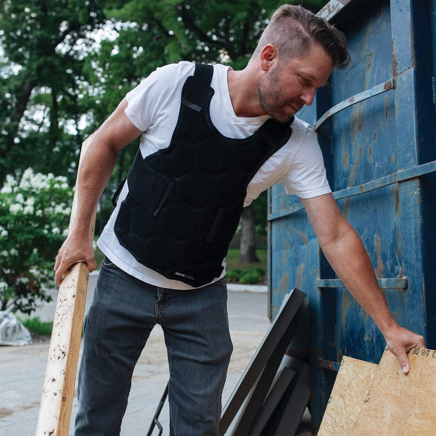 A person wearing the Xena Therapies Onyx Cool Safety Pro Vest lifts wooden boards by a blue metal container outdoors.