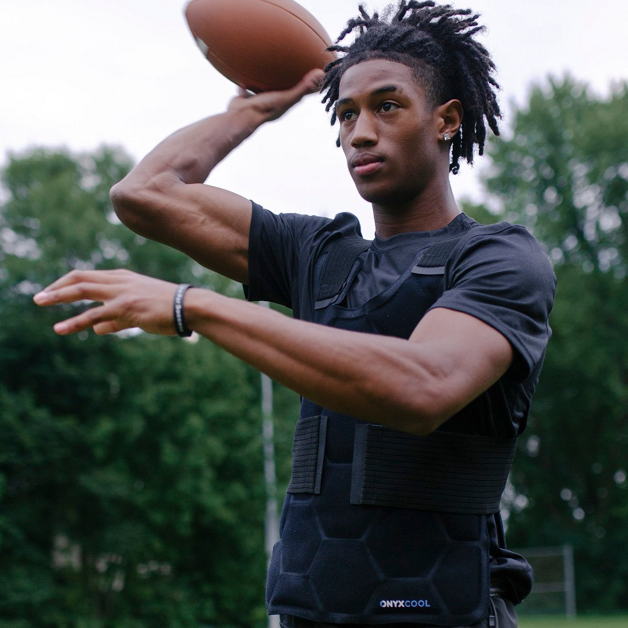 A person wears a Xena Therapies Onyx Cool Safety Pro Vest outdoors while holding a football. Trees are visible in the background.