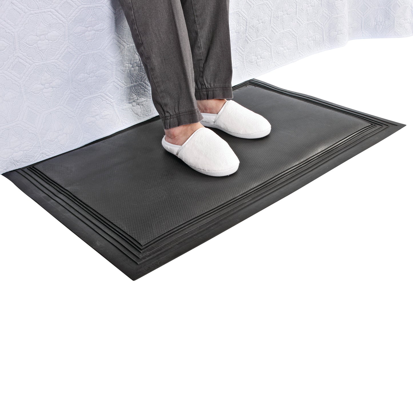 A person wearing white slippers stands on an AliMed Nonslip Exit Alarm Mat beside a bed with a white quilted cover.