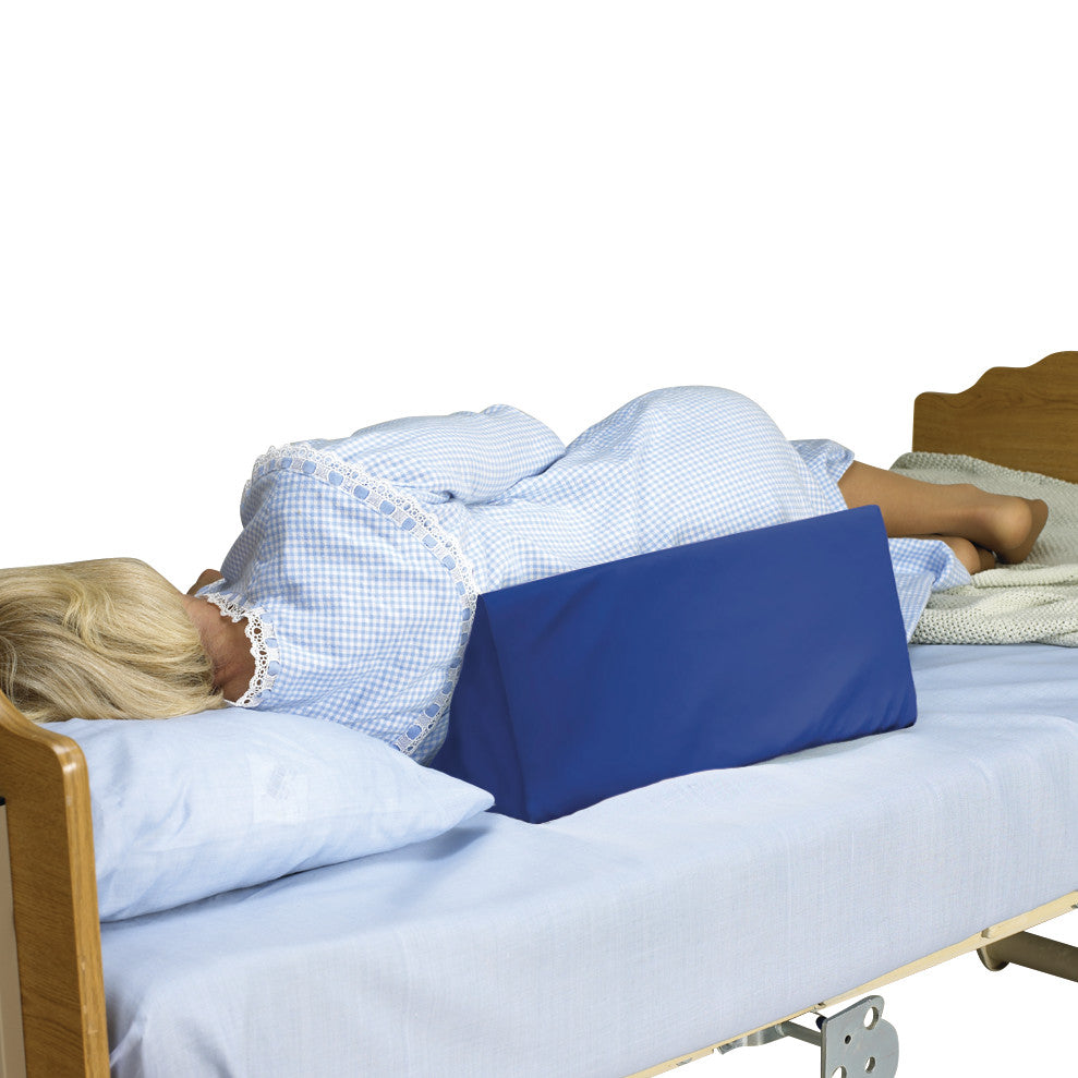 A person lies on a bed using the SkiL-Care Positioning Wedge with Cover for support, wearing a blue, checkered nightgown.