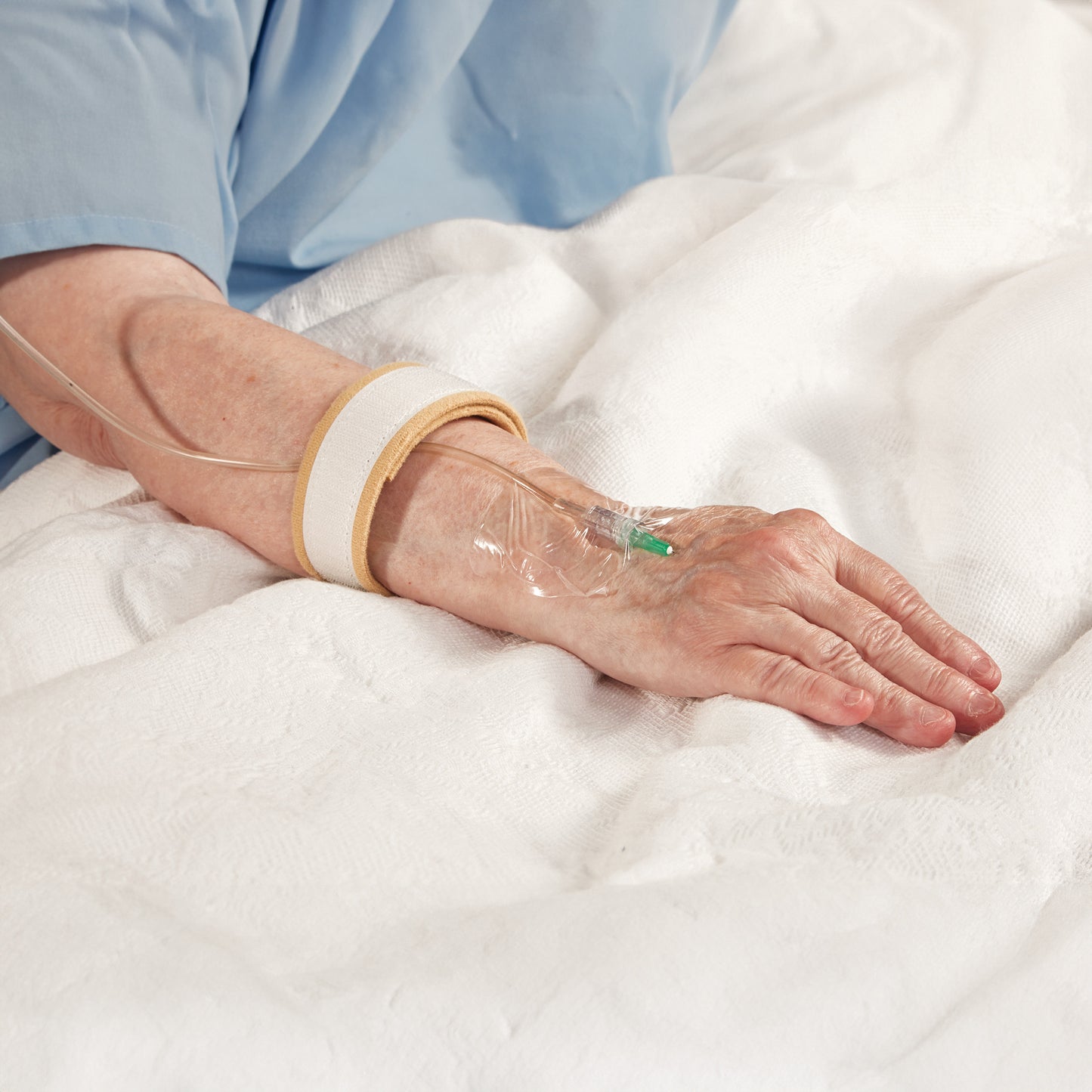 An elderly patient with an IV is resting. The AliMed AliStrap Soft Positioning Strap secures their arm on a white bed.