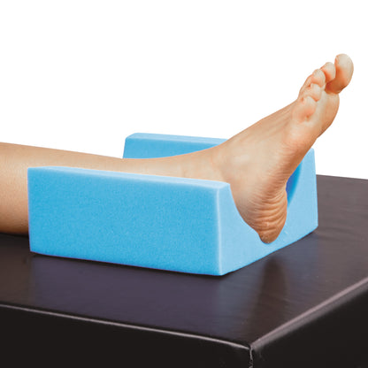 A foot rests on the AliMed Head/Heel Rest Positioner, a blue foam cushion, on a dark surface for support and elevation.